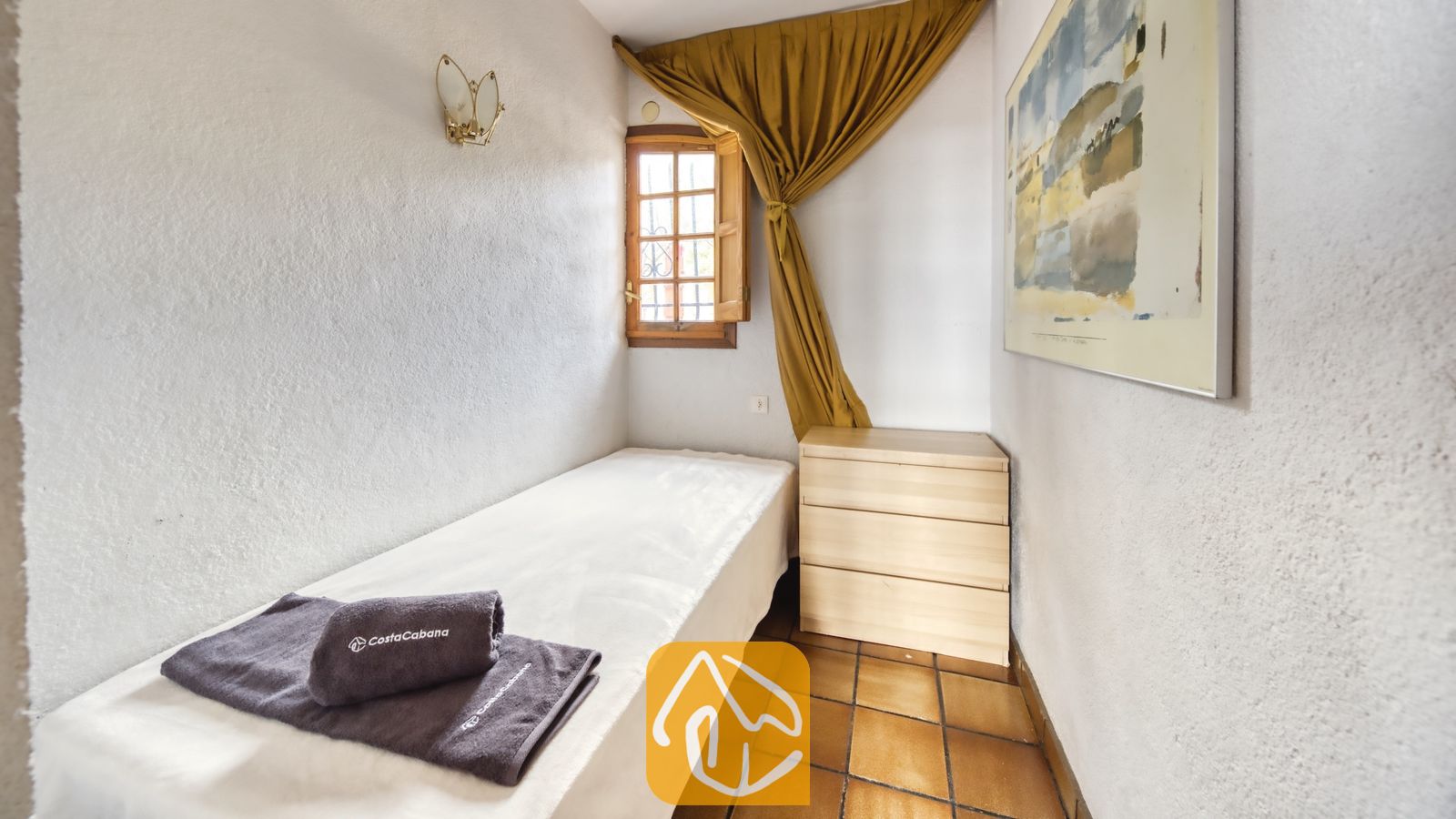 Vakantievilla's Costa Brava Spanje - Villa Janet - Een eenpersoonsbed met witte lakens en opgerolde grijskleurige handdoeken in een knusse slaapkamer voorzien van terracotta plavuizen en een klein houten raam.