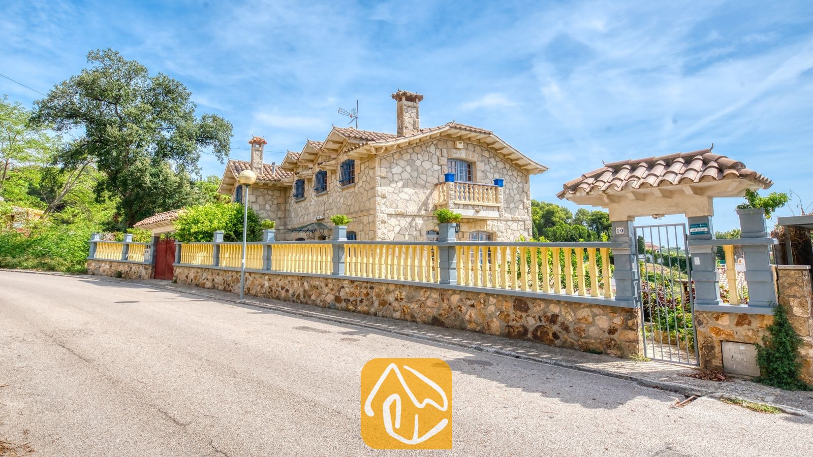 Vakantievilla's Costa Brava Spanje - Villa Janet - Buitenaanzicht van Villa Janet met de stenen gevel, pannendak en een geel-grijze omheining langs de weg.