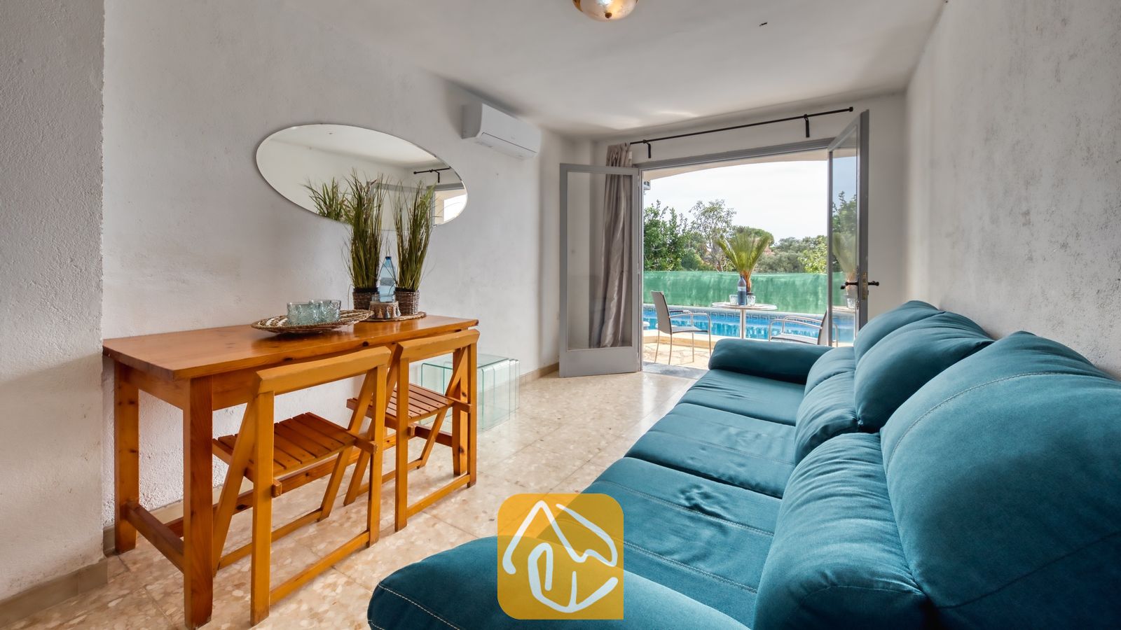 Vakantievilla's Costa Brava Spanje - Villa Janet - Woonkamer met een turquoise bank, een houten eettafel met twee klapstoelen en een ovale wandspiegel, met openslaande deuren naar een zwembadterras.
