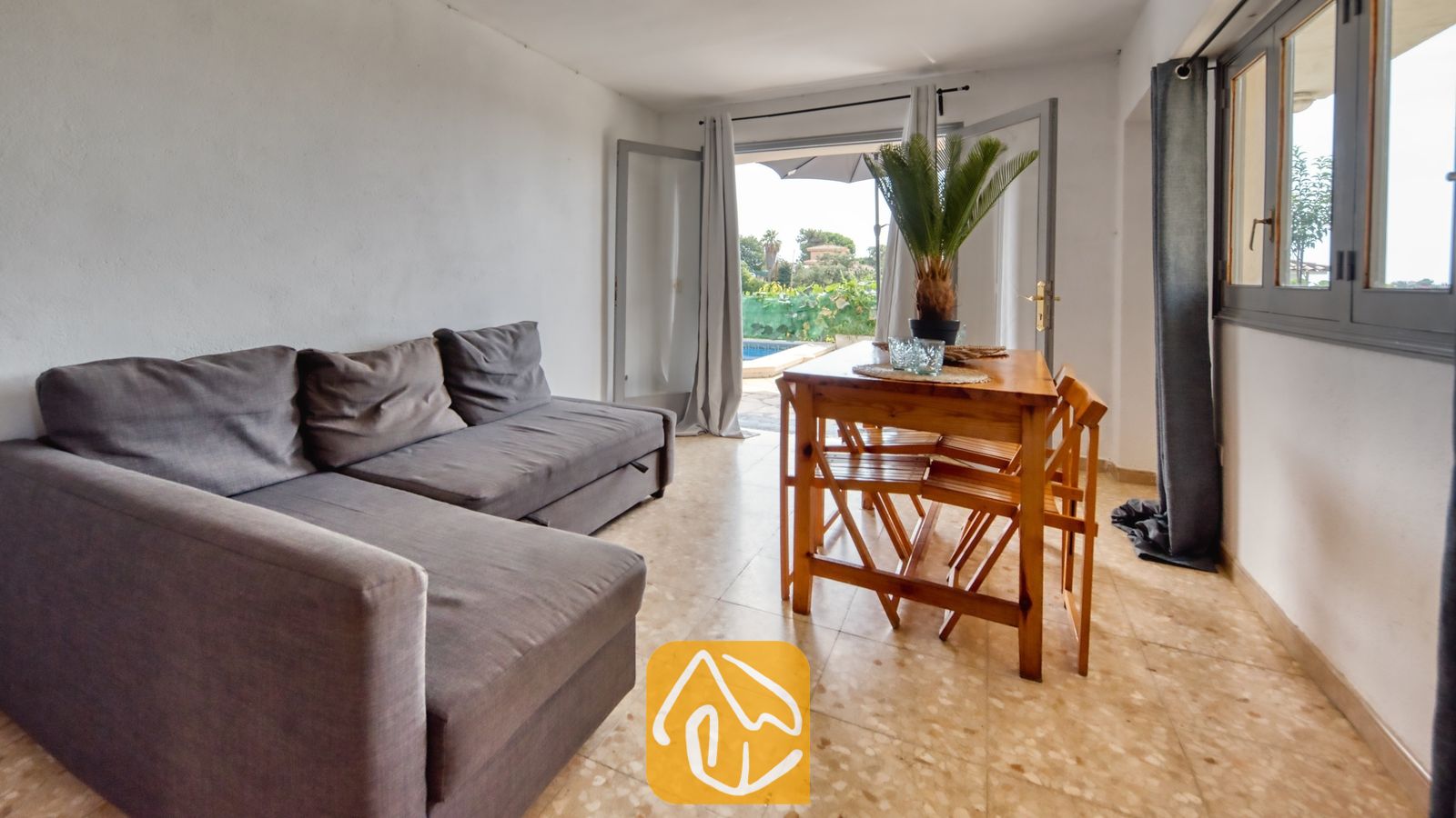 Vakantievilla's Costa Brava Spanje - Villa Janet - Woonkamer in Villa Janet met een grijze hoekbank, een houten eettafel met stoelen en open glazen deuren die leiden naar een buitenzwembad.