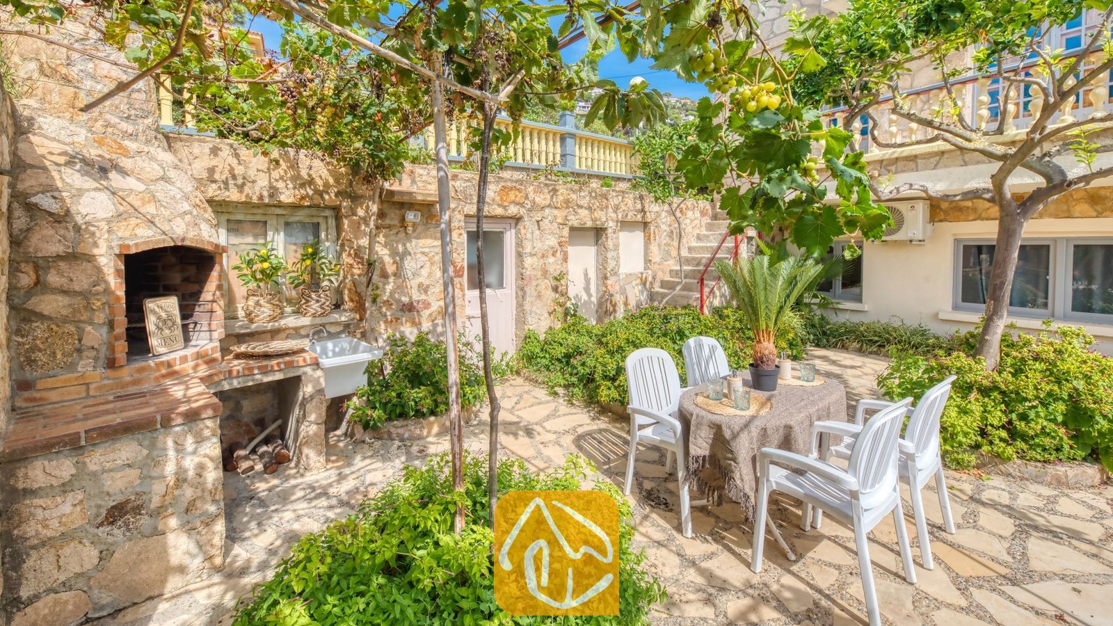 Vakantievilla's Costa Brava Spanje - Villa Janet - Stenen terras bij Villa Janet, voorzien van een ingebouwde stenen barbecue, een eettafel voor buiten met vier witte plastic stoelen en druivenranken erboven.