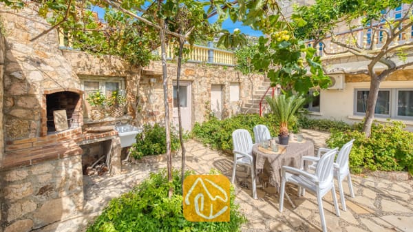 Vakantievilla's Costa Brava Spanje - Villa Janet - Stenen terras bij Villa Janet, voorzien van een ingebouwde stenen barbecue, een eettafel voor buiten met vier witte plastic stoelen en druivenranken erboven.