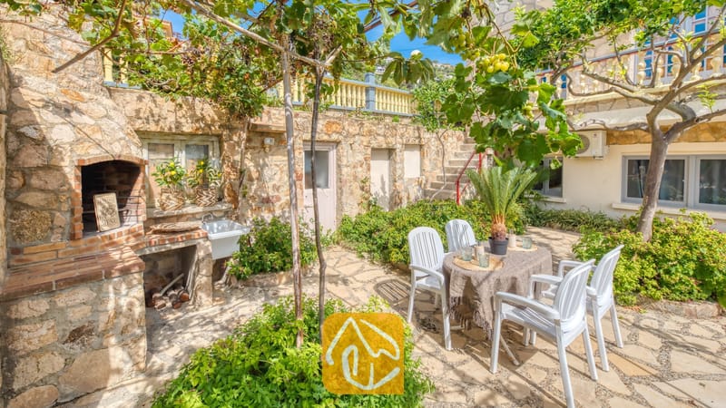 Vakantievilla's Costa Brava Spanje - Villa Janet - Stenen terras bij Villa Janet, voorzien van een ingebouwde stenen barbecue, een eettafel voor buiten met vier witte plastic stoelen en druivenranken erboven.