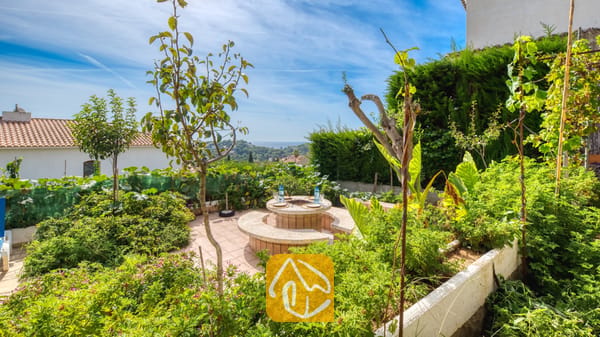 Vakantievilla's Costa Brava Spanje - Villa Janet - Buiteniterras van Villa Janet met een ronde stenen tafel en ingebouwde zitplaatsen, omringd door weelderige groene plantenbakken, kleine bomen en een ver uitzicht op zee onder een blauwe hemel.