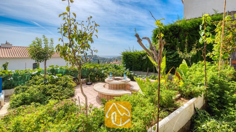 Vakantievilla's Costa Brava Spanje - Villa Janet - Buiteniterras van Villa Janet met een ronde stenen tafel en ingebouwde zitplaatsen, omringd door weelderige groene plantenbakken, kleine bomen en een ver uitzicht op zee onder een blauwe hemel.