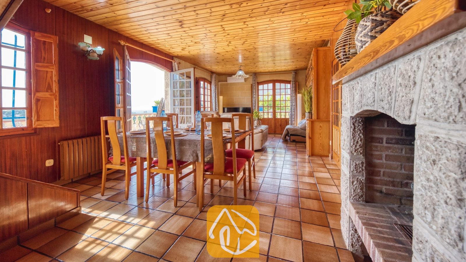 Vakantievilla's Costa Brava Spanje - Villa Janet - Houten eettafel met stoelen met rode kussens binnen in Villa Janet, voorzien van tegelvloeren, houten lambrisering op muren en plafond, en een grote stenen open haard.