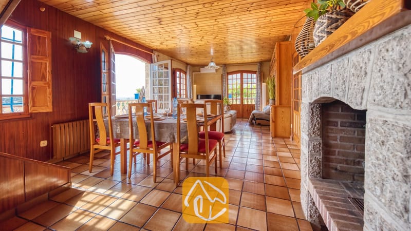 Vakantievilla's Costa Brava Spanje - Villa Janet - Houten eettafel met stoelen met rode kussens binnen in Villa Janet, voorzien van tegelvloeren, houten lambrisering op muren en plafond, en een grote stenen open haard.