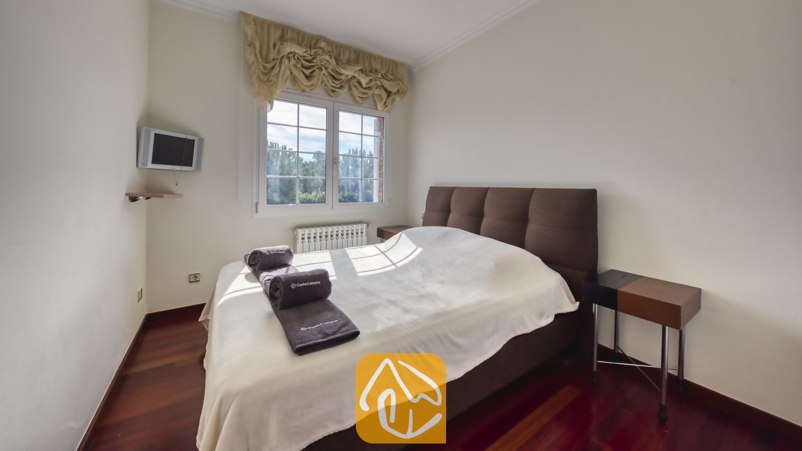 Vakantievilla's Costa Brava Spanje - Villa Beyonce - Slaapkamer met een bruin gestoffeerd tweepersoonsbed, opgevouwen grijze handdoeken, wit beddengoed en twee houten nachtkastjes op een donkere houten vloer.