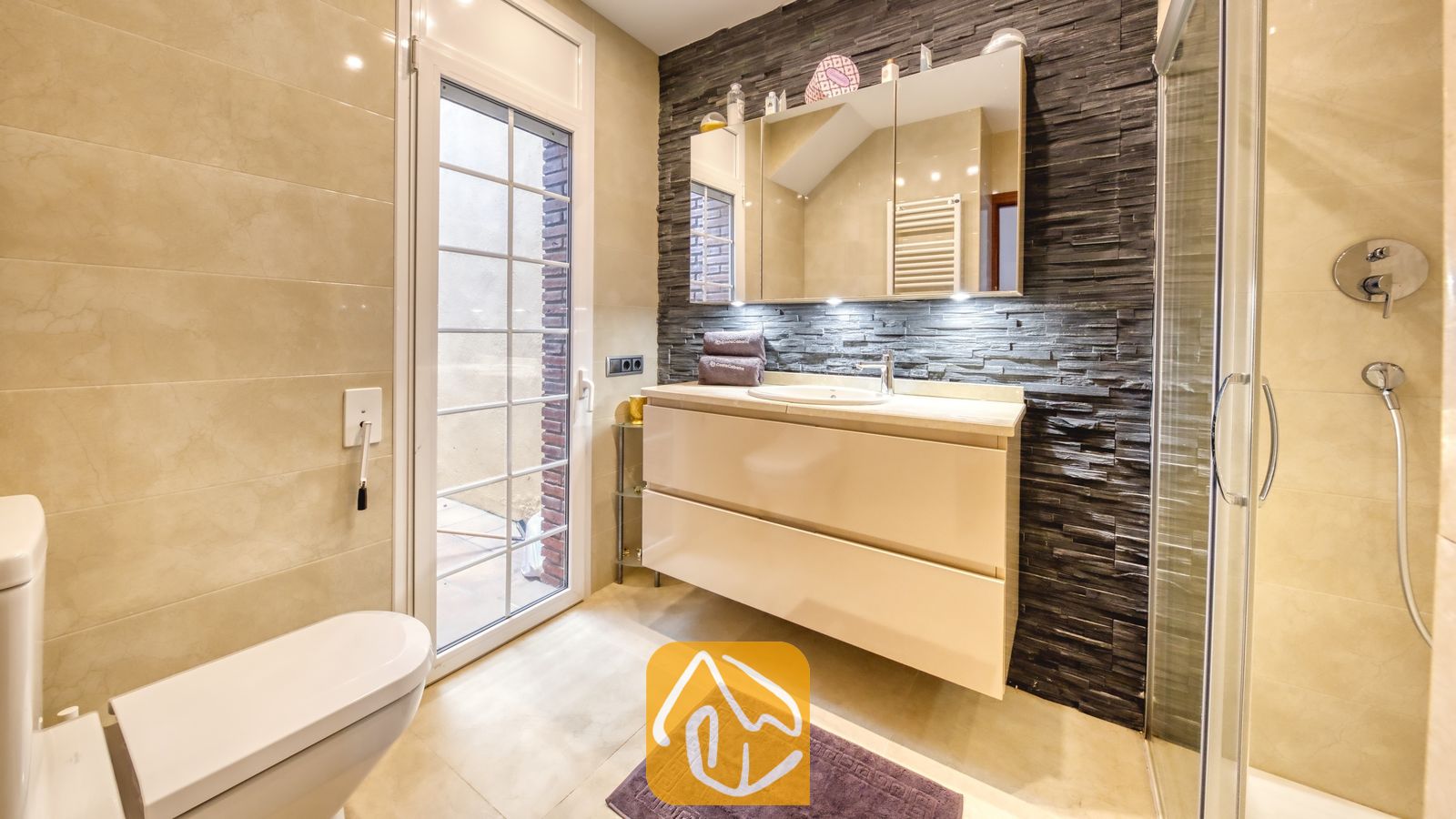 Vakantievilla's Costa Brava Spanje - Villa Beyonce - Moderne badkamer met een crème badkamermeubel en een opzetwastafel tegen een donkere, getextureerde stenen wand, een glazen deur naar buiten en een spiegelkast.