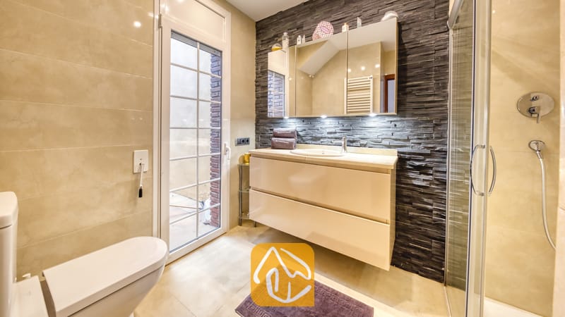Villas de vacances Costa Brava Espagne - Villa Beyonce - Salle de bains moderne comprenant un meuble sous-vasque crème surmonté d'une vasque à poser adossé à un mur en pierre texturée foncée, une porte vitrée donnant sur l'extérieur et une armoire de toilette miroir.