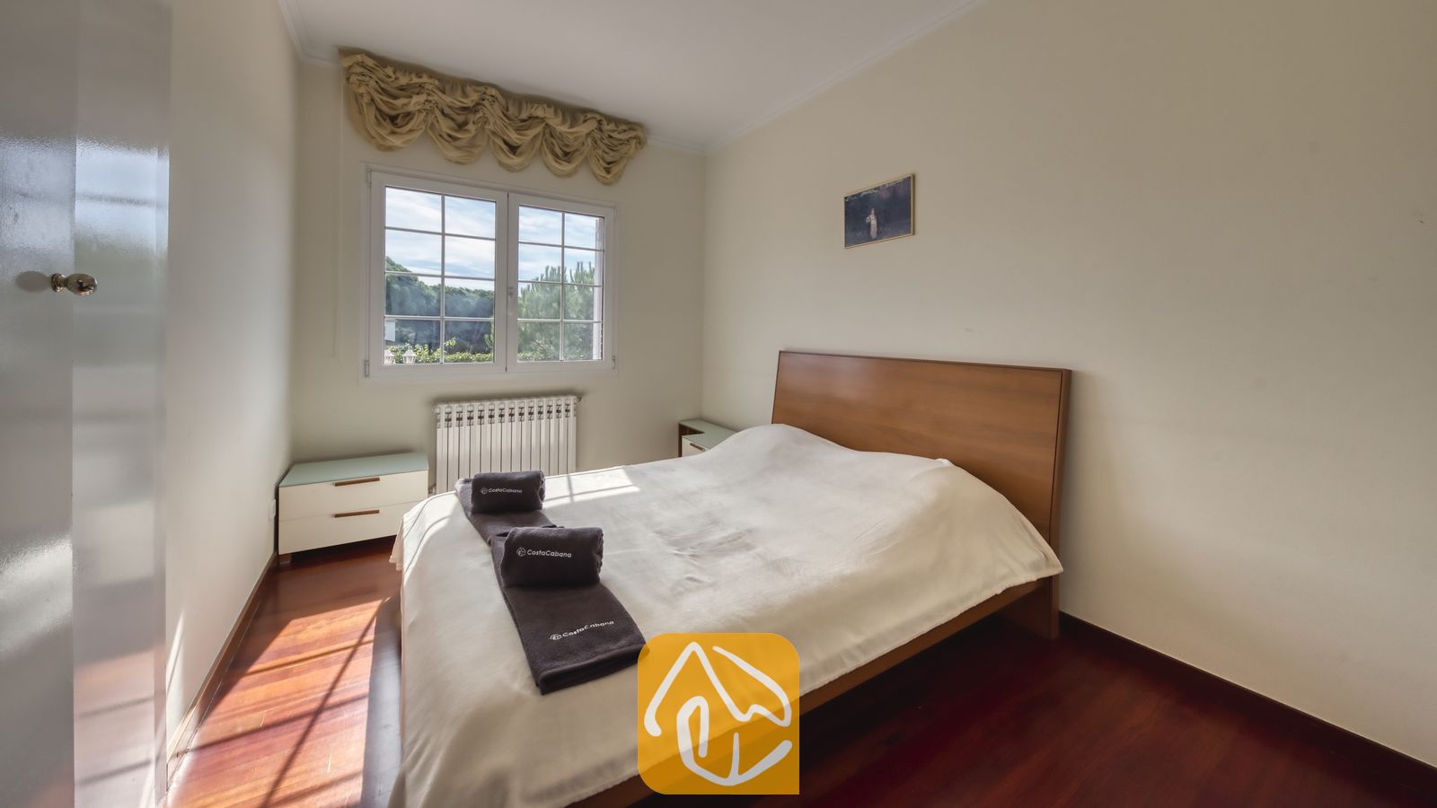 Vakantievilla's Costa Brava Spanje - Villa Beyonce - Slaapkamer met een houten bedframe, donkere opgerolde handdoeken op een wit bed en een raam met gefranjerde gordijntjes.
