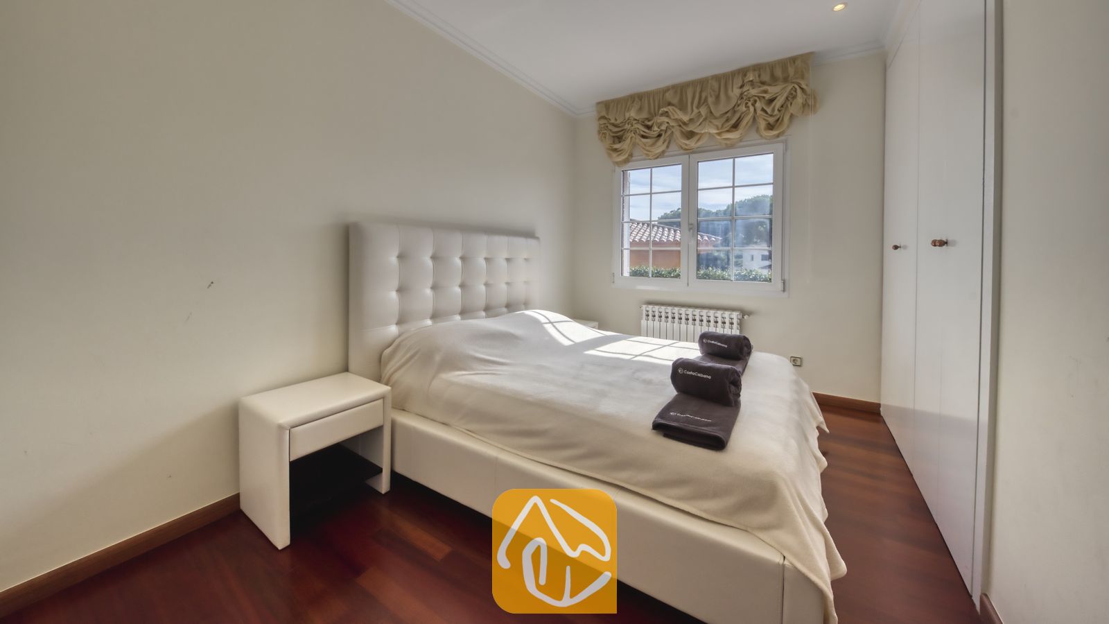 Vakantievilla's Costa Brava Spanje - Villa Beyonce - Slaapkamer met een wit capitoneerlederen bed, donkere houten vloeren, een wit nachtkastje, opgerolde donkere handdoeken en een raam met gegolfde beige gordijnen.