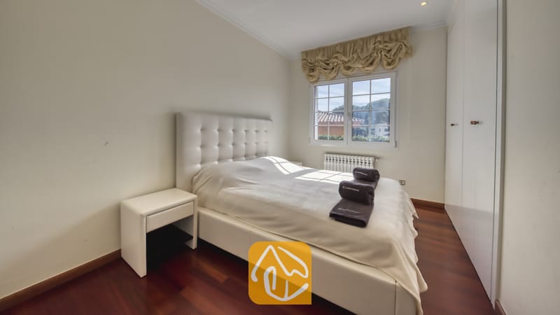 Villas de vacances Costa Brava Espagne - Villa Beyonce - Chambre avec un lit capitonné en cuir blanc, des planchers en bois foncé, une table de chevet blanche, des serviettes foncées enroulées et une fenêtre habillée de rideaux beige à volants.