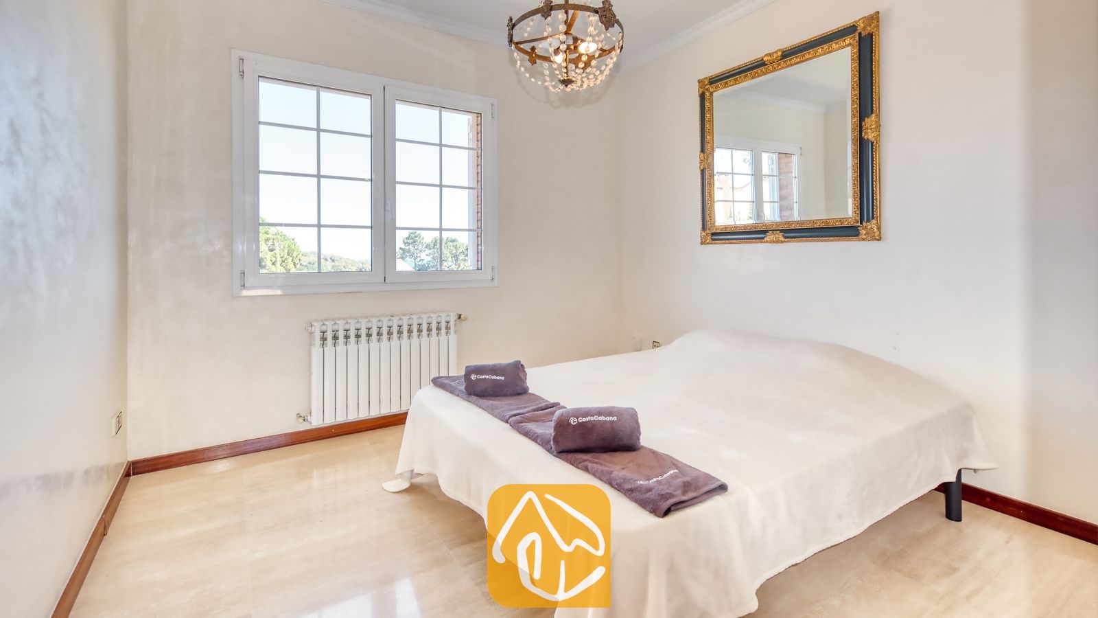 Vakantievilla's Costa Brava Spanje - Villa Beyonce - Slaapkamer met een tweepersoonsbed opgemaakt in wit linnen, opgevouwen grijze handdoeken, een grote, sierlijk ingelijste spiegel aan de muur en een kristallen kroonluchter met meerdere lampjes.