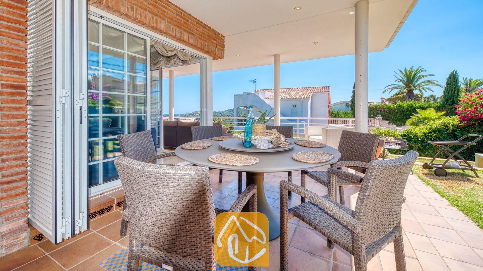 Vakantievilla's Costa Brava Spanje - Villa Beyonce - Overdekt buitenterras om buiten te dineren bij Villa Beyonce, voorzien van een ronde tafel met geweven rieten stoelen, tegelvloeren en glazen schuifdeuren die naar binnen leiden.