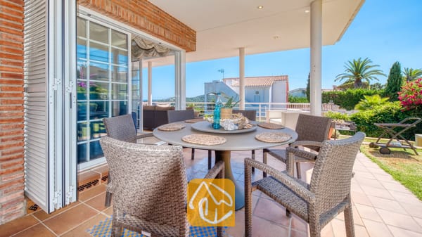 Vakantievilla's Costa Brava Spanje - Villa Beyonce - Overdekt buitenterras om buiten te dineren bij Villa Beyonce, voorzien van een ronde tafel met geweven rieten stoelen, tegelvloeren en glazen schuifdeuren die naar binnen leiden.