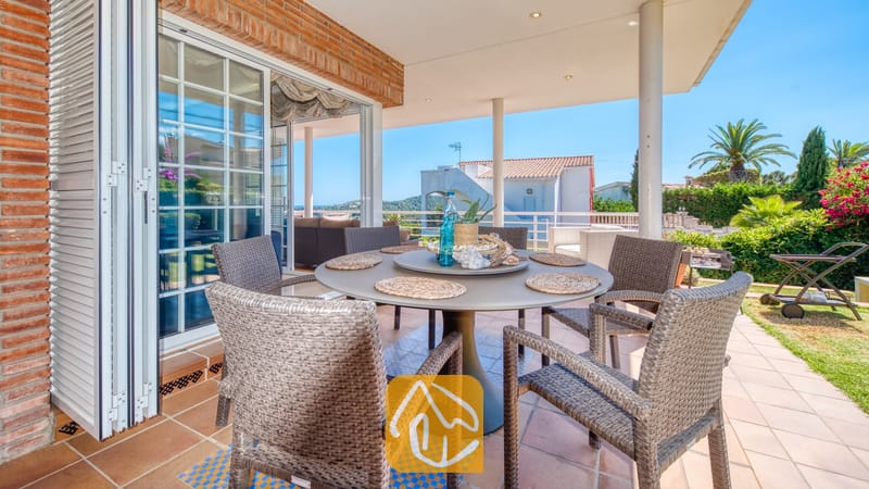 Vakantievilla's Costa Brava Spanje - Villa Beyonce - Overdekt buitenterras om buiten te dineren bij Villa Beyonce, voorzien van een ronde tafel met geweven rieten stoelen, tegelvloeren en glazen schuifdeuren die naar binnen leiden.