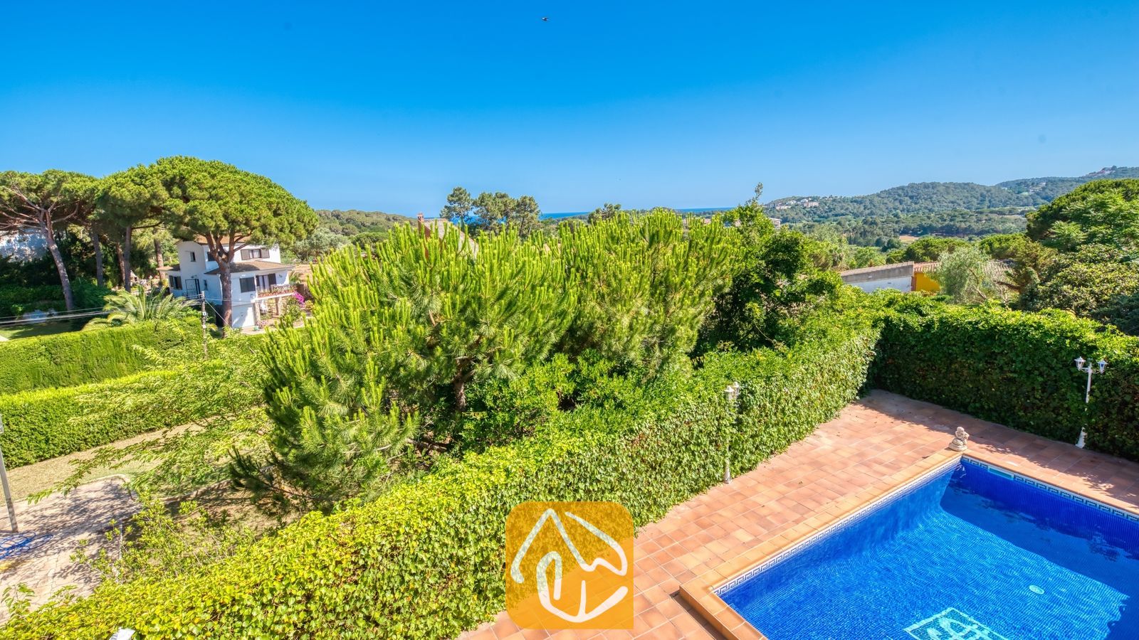 Vakantievilla's Costa Brava Spanje - Villa Beyonce - Uitzicht naar buiten vanuit Villa Beyonce met een zwembad omringd door terracotta tegels, groene hagen en bomen in de verte onder een helderblauwe hemel.