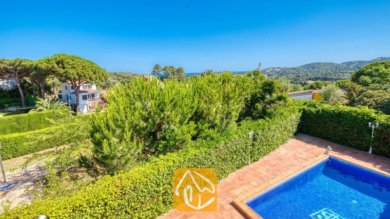 Villas de vacances Costa Brava Espagne - Villa Beyonce - Vue extérieure de la Villa Beyoncé montrant une piscine entourée de carrelage en terre cuite, de hauts hedges verts et d'arbres au loin sous un ciel bleu dégagé.