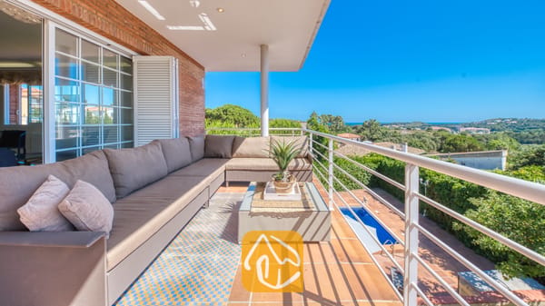 Vakantievilla's Costa Brava Spanje - Villa Beyonce - Overdekt balkon bij Villa Beyonce met een grote hoekbank, een salontafel met glazen blad en blauw-met-gele tegelvloer, met uitzicht op de bomen en een zwembad.