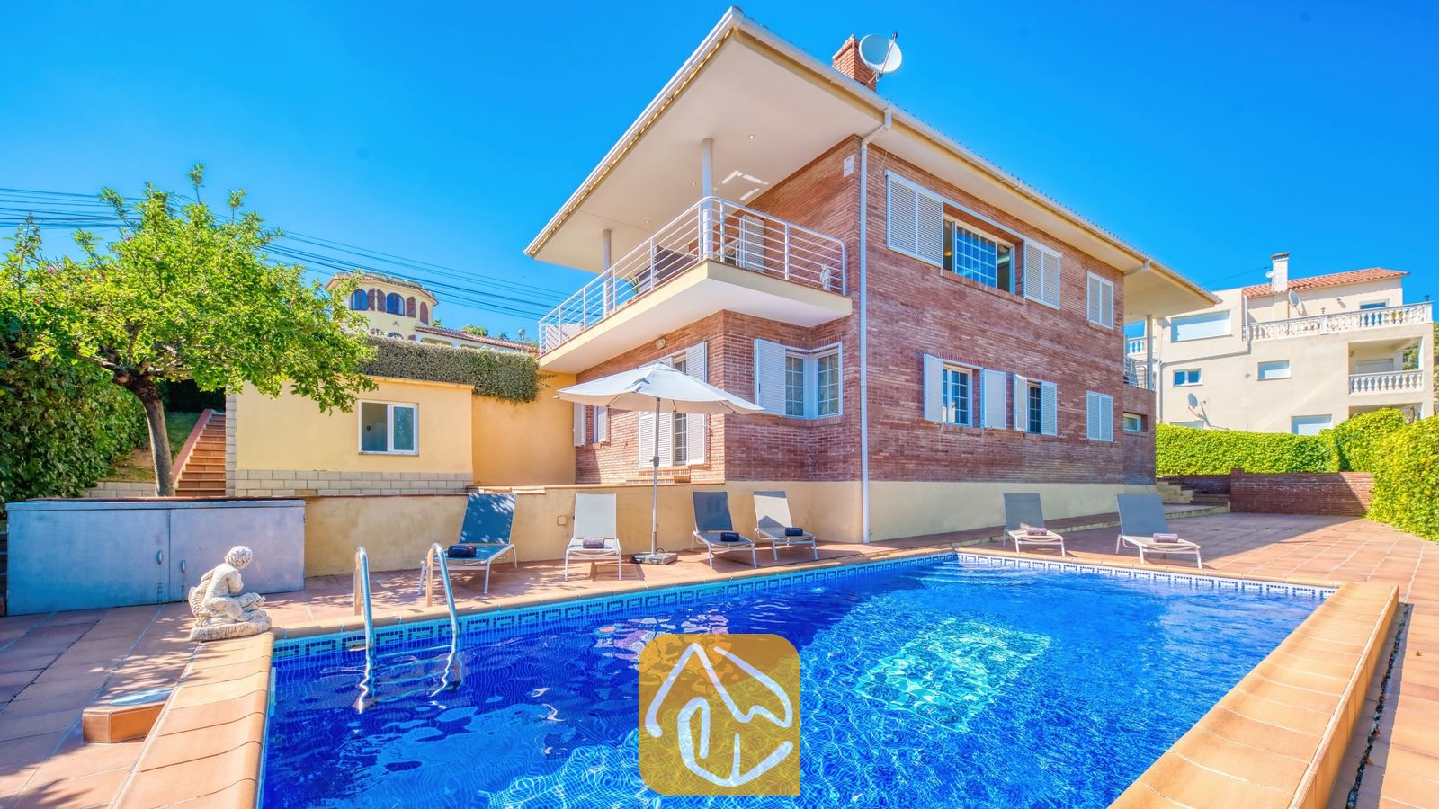 Vakantievilla's Costa Brava Spanje - Villa Beyonce - Buitenaanzicht van Villa Beyonce met een rechthoekig zwembad met blauwe tegels, ligstoelen en een stenen gebouw van twee verdiepingen onder een helderblauwe hemel