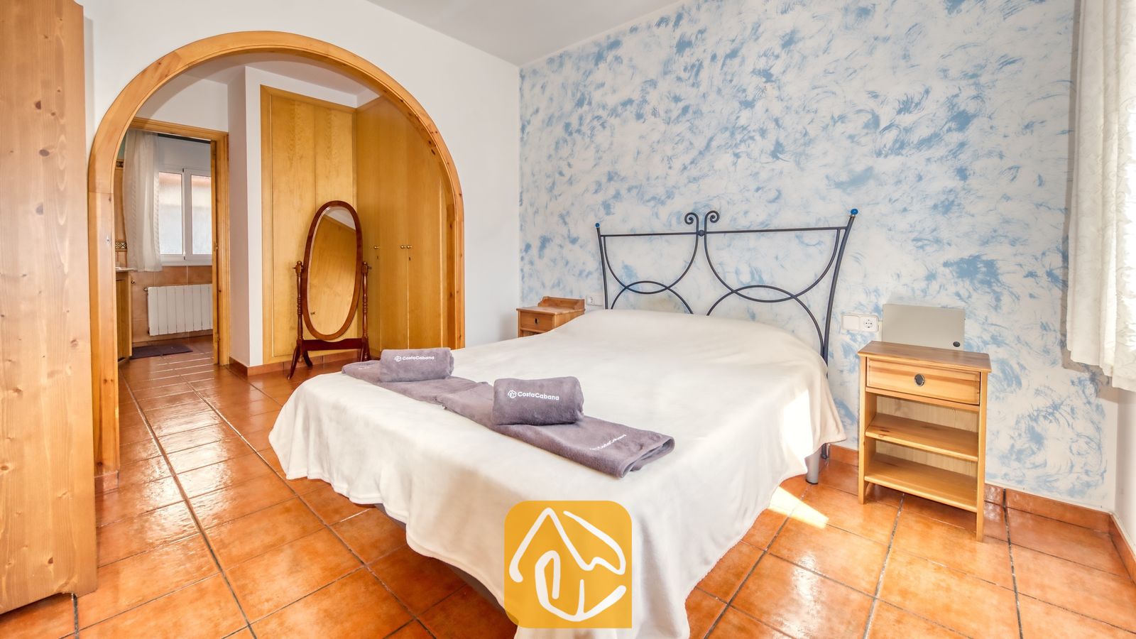 Vakantievilla's Costa Brava Spanje - Villa Valeria - Slaapkamer met een tweepersoonsbed, een metalen hoofdeinde en een blauwe accentmuur met patroon, voorzien van terracotta plavuizen en een houten toog die leidt naar een aangrenzende kamer.