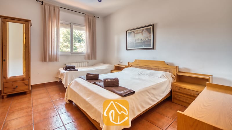 Holiday villas Costa Brava Spain - Villa Valeria - Bedroom with a double bed, a single bed, wooden furniture, tiled floors, and a window with curtains at Villa Valeria.