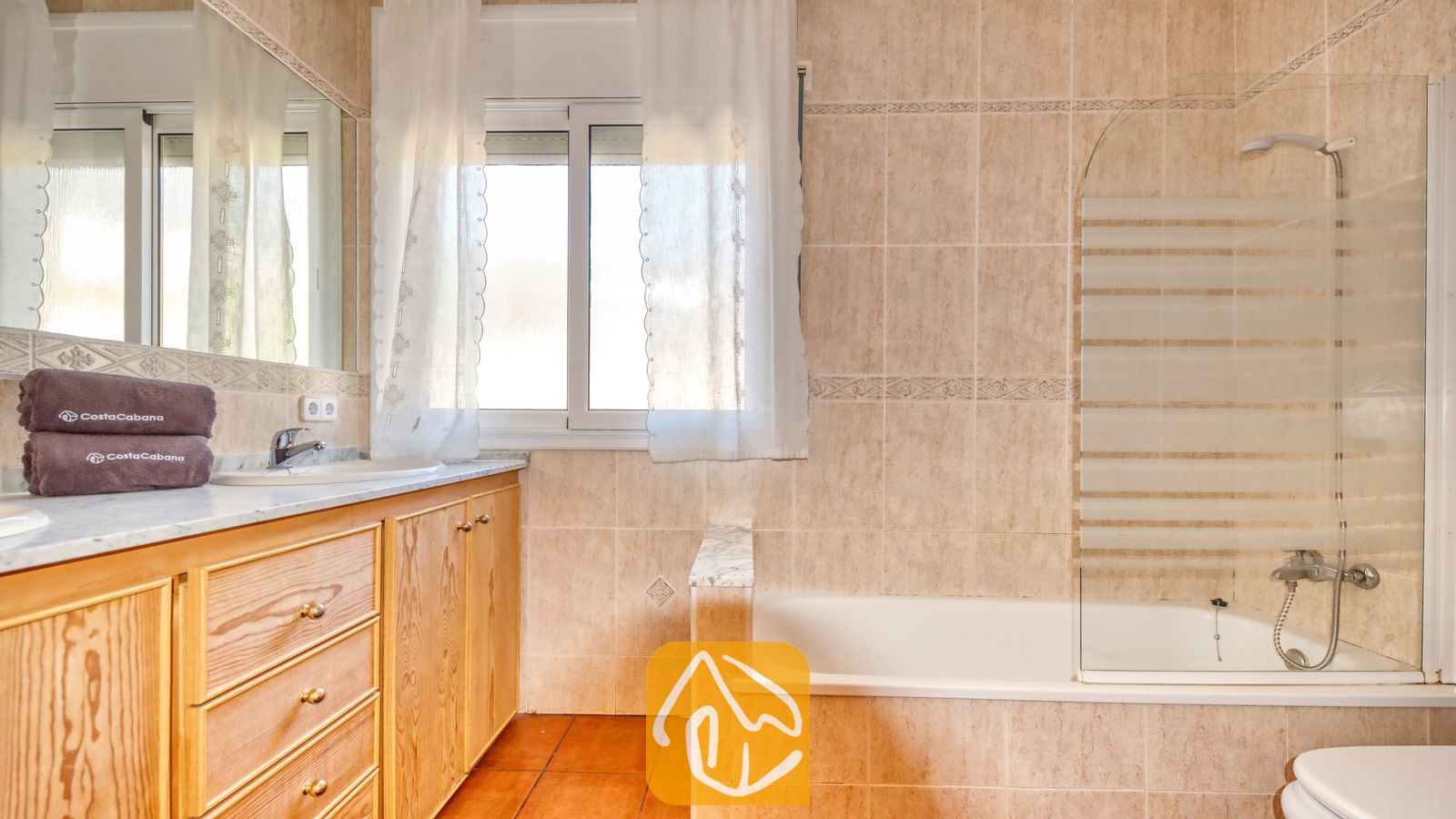 Vakantievilla's Costa Brava Spanje - Villa Valeria - Badkamer met beige betegelde muren, een houten badkamermeubel met lades, een wit bad met een glazen scherm en een raam met inbetween-gordijnen.