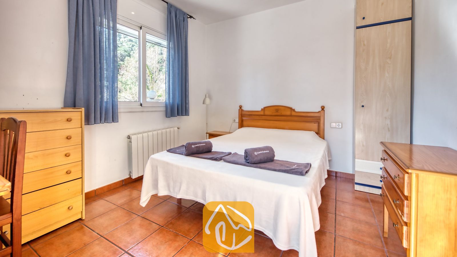 Vakantievilla's Costa Brava Spanje - Villa Valeria - Slaapkamer met een houten tweepersoonsbed, twee opgevouwen grijze handdoeken op het witte beddengoed, houten dressoirs en blauwe gordijnen die het raam omlijsten.