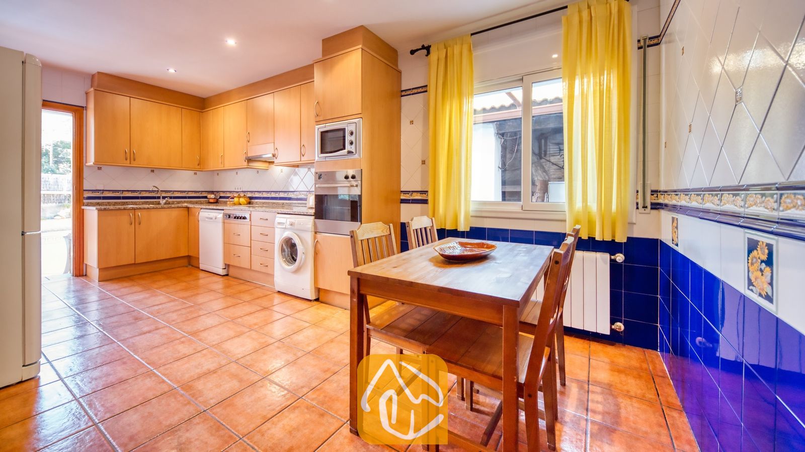 Vakantievilla's Costa Brava Spanje - Villa Valeria - Keuken met houten kasten, een eettafel met vier stoelen, tegelvloer en gele gordijnen voor het raam.