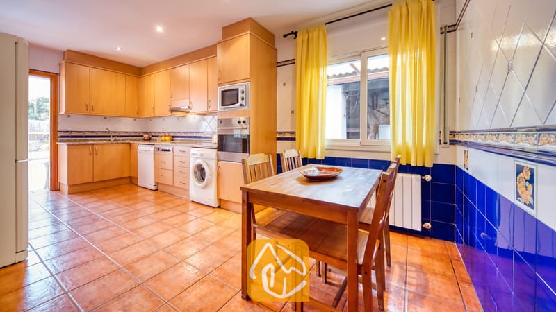 Vakantievilla's Costa Brava Spanje - Villa Valeria - Keuken met houten kasten, een eettafel met vier stoelen, tegelvloer en gele gordijnen voor het raam.
