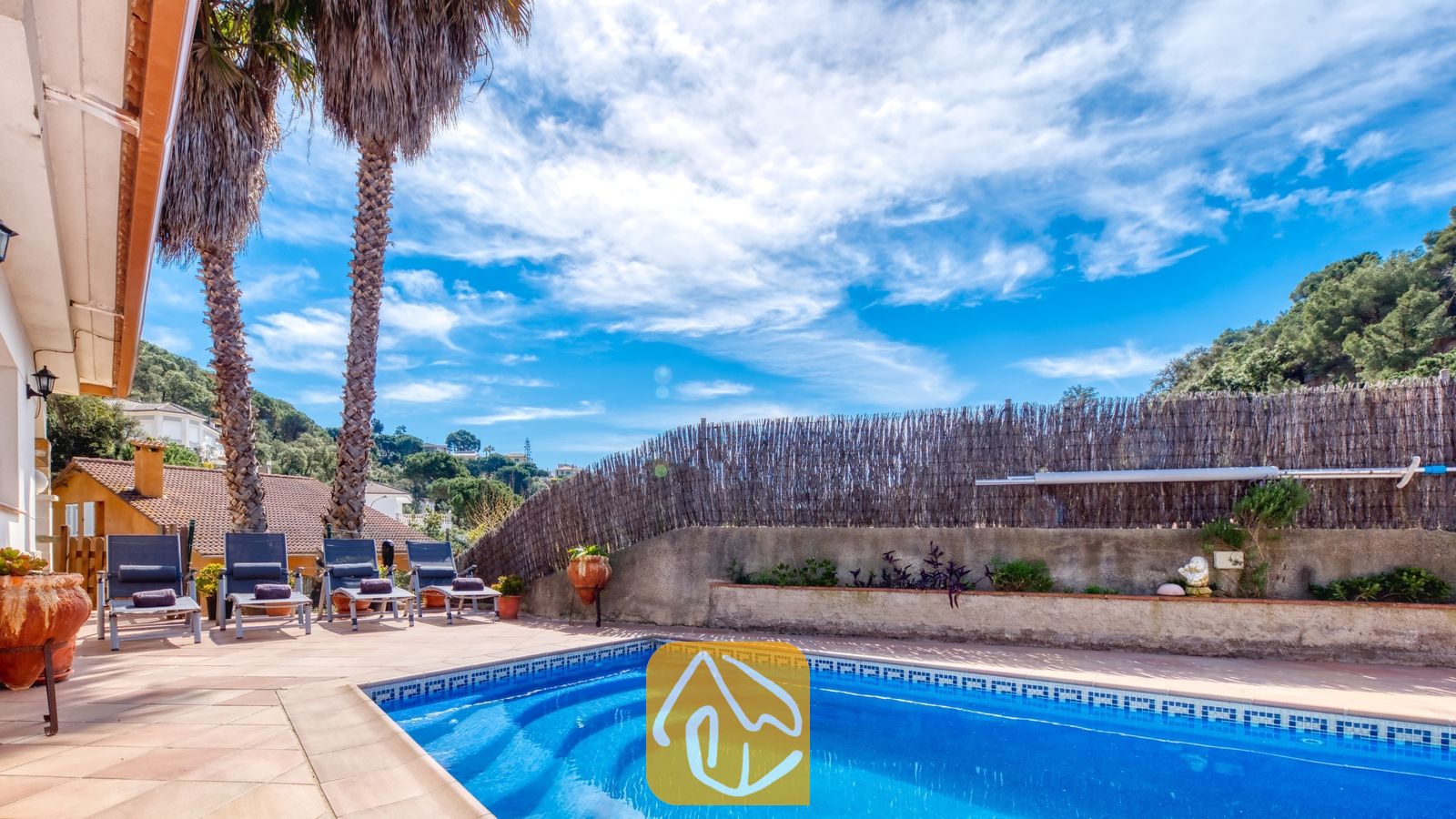 Vakantievilla's Costa Brava Spanje - Villa Valeria - Privé buitenzwembad bij Villa Valeria met helderblauw water, een betegelde rand, ligstoelen en hoge palmbomen tegen een bewolkte blauwe lucht.