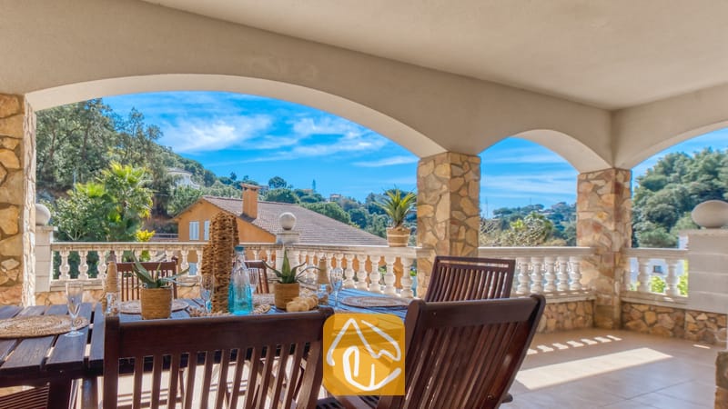 Vakantievilla's Costa Brava Spanje - Villa Valeria - Overdekt terras bij Villa Valeria, voorzien van een houten eettafel met servies, stenen pilaren en een boogvormig uitzicht op de groene heuvels.