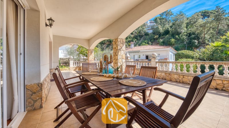 Vakantievilla's Costa Brava Spanje - Villa Valeria - Overdekt terras van Villa Valeria, voorzien van een donkerhouten eettafel gedekt met geweven placemats, omringd door bijpassende stoelen en met uitzicht op een met bomen begroeide heuvel.