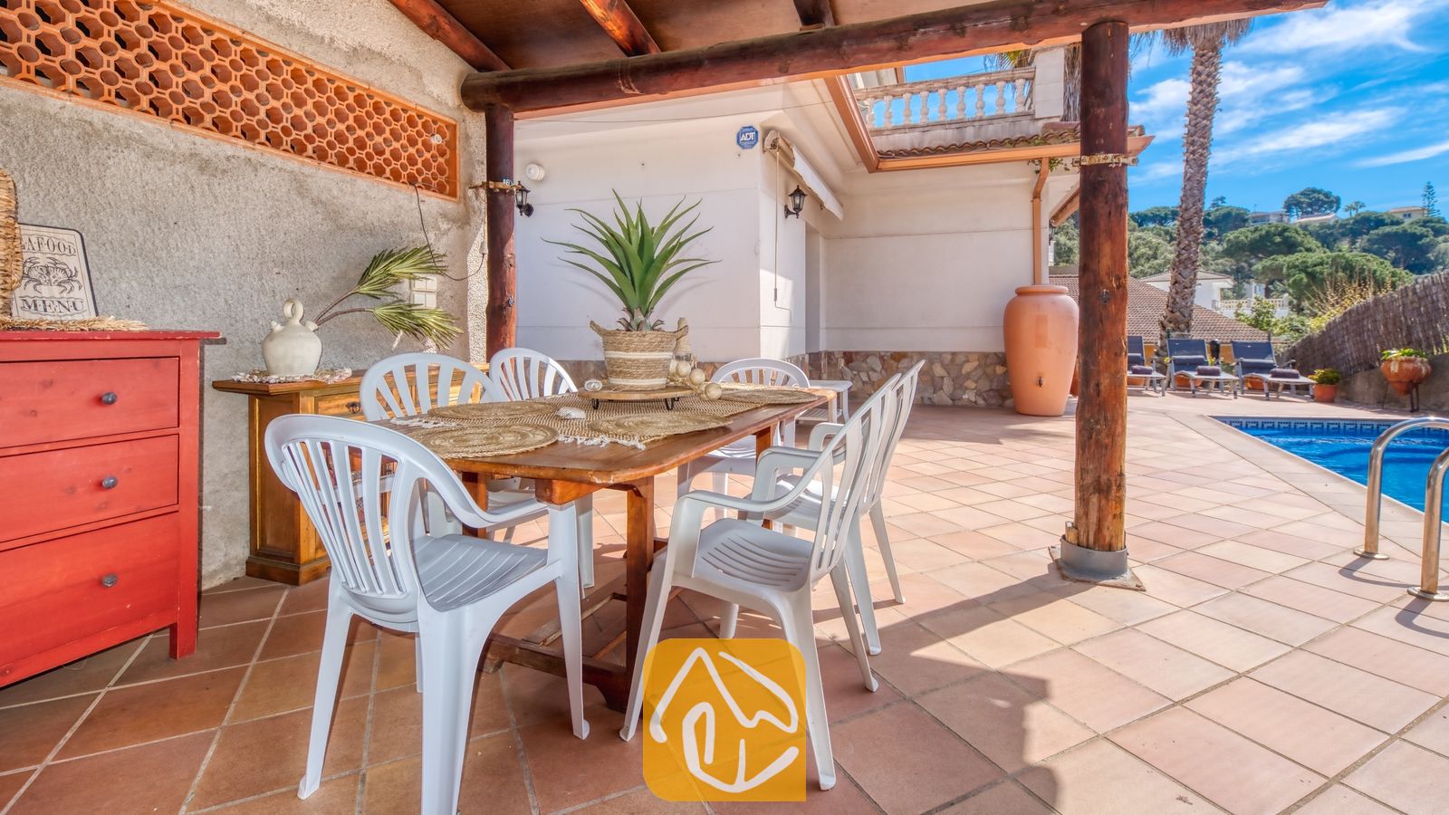Vakantievilla's Costa Brava Spanje - Villa Valeria - Overdekt buitenterras bij Villa Valeria met een houten tafel, witte kunststof stoelen en een plant als middelpunt, gelegen naast een betegeld zwembad.