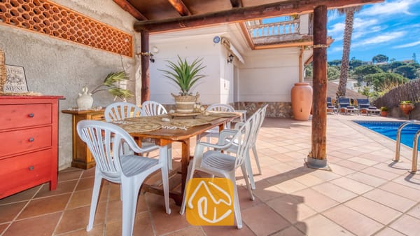 Vakantievilla's Costa Brava Spanje - Villa Valeria - Overdekt buitenterras bij Villa Valeria met een houten tafel, witte kunststof stoelen en een plant als middelpunt, gelegen naast een betegeld zwembad.