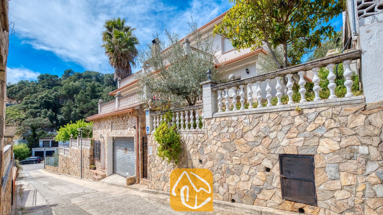 Vakantievilla's Costa Brava Spanje - Villa Valeria - Buitenaanzicht van Villa Valeria in Spanje met een stenen keermuur met witte balustrades, een garagedeur en een palmboom onder een blauwe lucht.