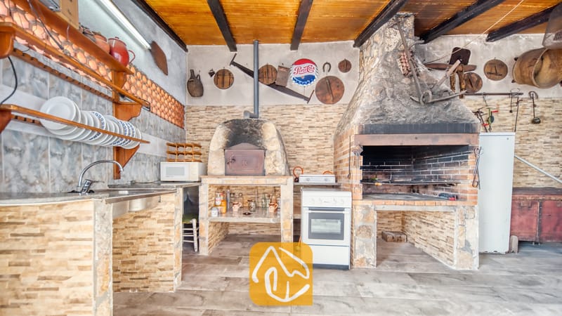 Vakantievilla's Costa Brava Spanje - Villa Valeria - Overdekte buitenkeuken bij Villa Valeria, voorzien van een stenen barbecue-openhaard, een stenen pizzaoven, een metalen spoelbak en rustieke wanddecoraties.