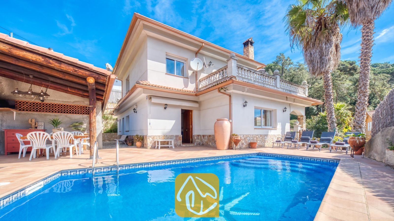 Vakantievilla's Costa Brava Spanje - Villa Valeria - Buitenaanzicht van Villa Valeria, met een zwembad voorzien van een tegelmozaïek, een witte villa van twee verdiepingen met stenen accenten, een overdekt terras met witte stoelen en hoge palmbomen.