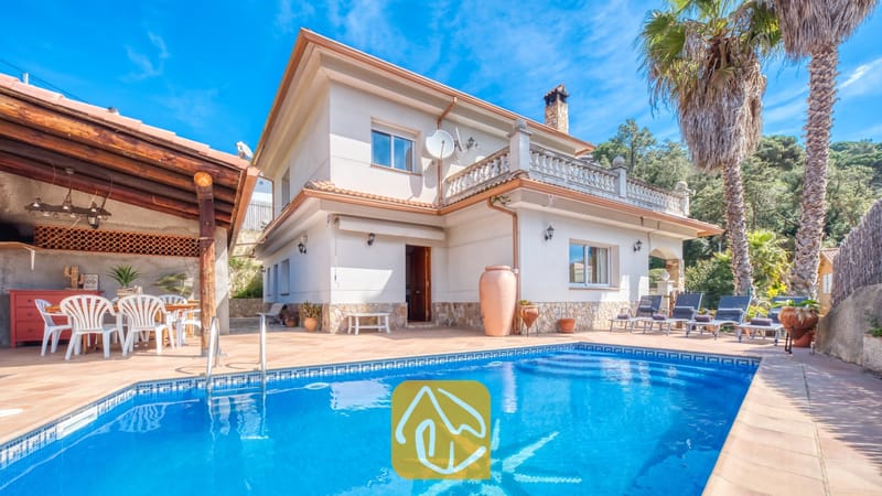 Vakantievilla's Costa Brava Spanje - Villa Valeria - Buitenaanzicht van Villa Valeria, met een zwembad voorzien van een tegelmozaïek, een witte villa van twee verdiepingen met stenen accenten, een overdekt terras met witte stoelen en hoge palmbomen.
