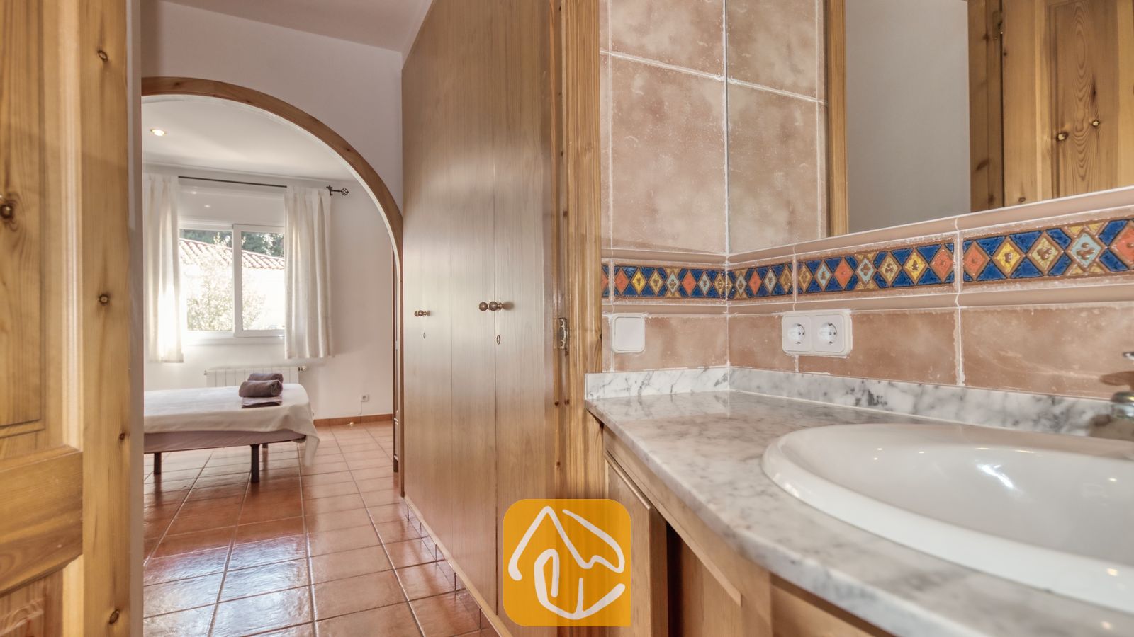 Vakantievilla's Costa Brava Spanje - Villa Valeria - Badkamermeubel met marmeren blad en ingebouwde wastafel, voorzien van beige tegels op de muren en een decoratieve blauw-oranje sierrand, dat via een houten toogdeur uitkomt in een slaapkamer met een win