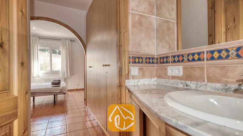 Holiday villas Costa Brava Spain - Villa Valeria - Bathroom vanity with marble countertop and inset sink, featuring beige tiled walls with a decorative blue and orange patterned border, opening through a wooden arched doorway into a bedroom with a win