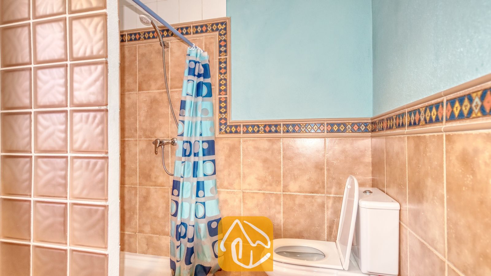 Vakantievilla's Costa Brava Spanje - Villa Valeria - Badkamer voorzien van beige betegelde muren met een sierlijke tegelrand, een blauw douchevordijn met geometrische patronen en een wit toilet naast een scheidingswand van glasstenen.