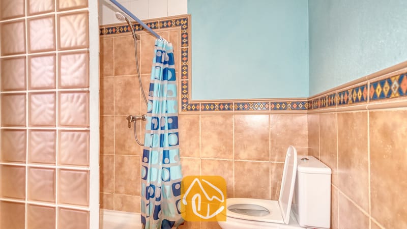 Holiday villas Costa Brava Spain - Villa Valeria - Bathroom featuring beige tiled walls with patterned border tiles, a blue shower curtain with geometric patterns, and a white toilet next to a glass block partition.