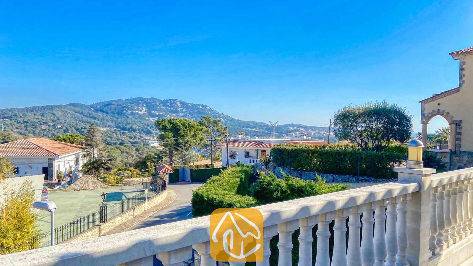 Vakantievilla's Costa Brava Spanje - Villa Primavera - Balkonterras van Villa Primavera met uitzicht op groene heuvels onder een helderblauwe lucht, met een verharde weg en naburige huizen beneden.