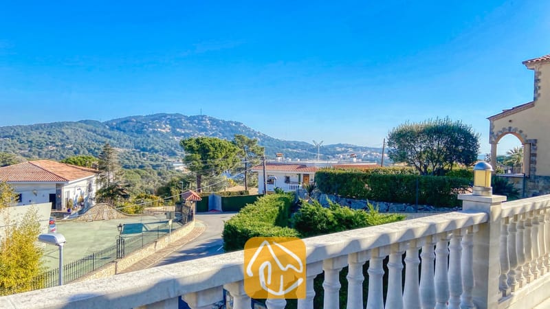Villas de vacances Costa Brava Espagne - Villa Primavera - Terrasse à balustrades de la Villa Primavera surplombant des collines verdoyantes sous un ciel bleu éclatant, avec une route pavée et des maisons voisines en contrebas.