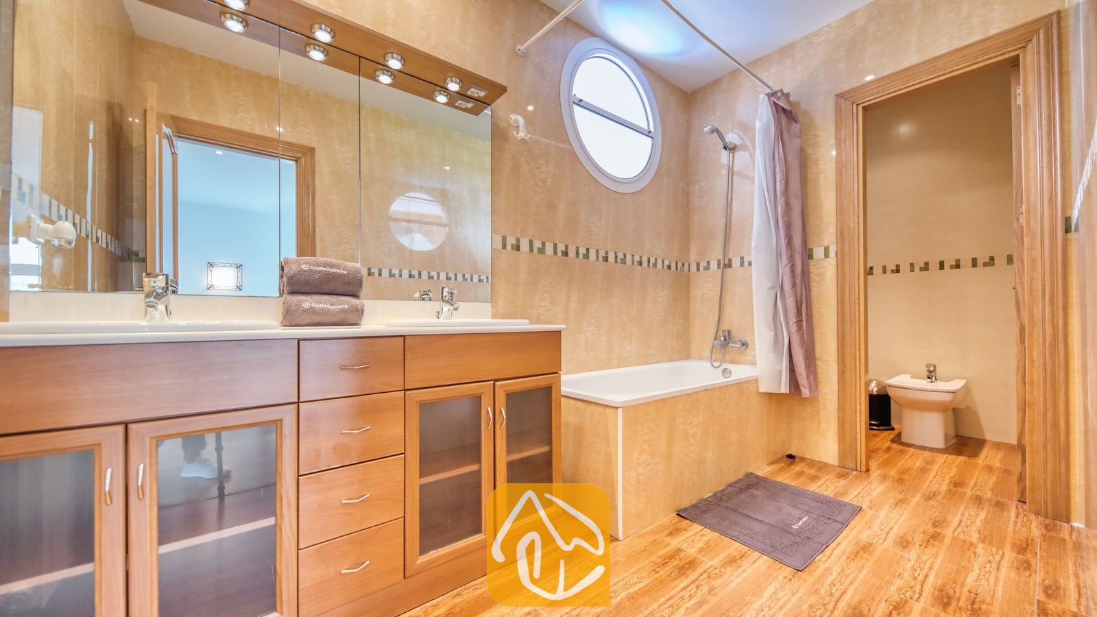 Vakantievilla's Costa Brava Spanje - Villa Primavera - Badkamer voorzien van een houten dubbele wastafel met een grote spiegel, een ligbad met een douchesnoer onder een rond raam, en betegelde muren en vloeren.