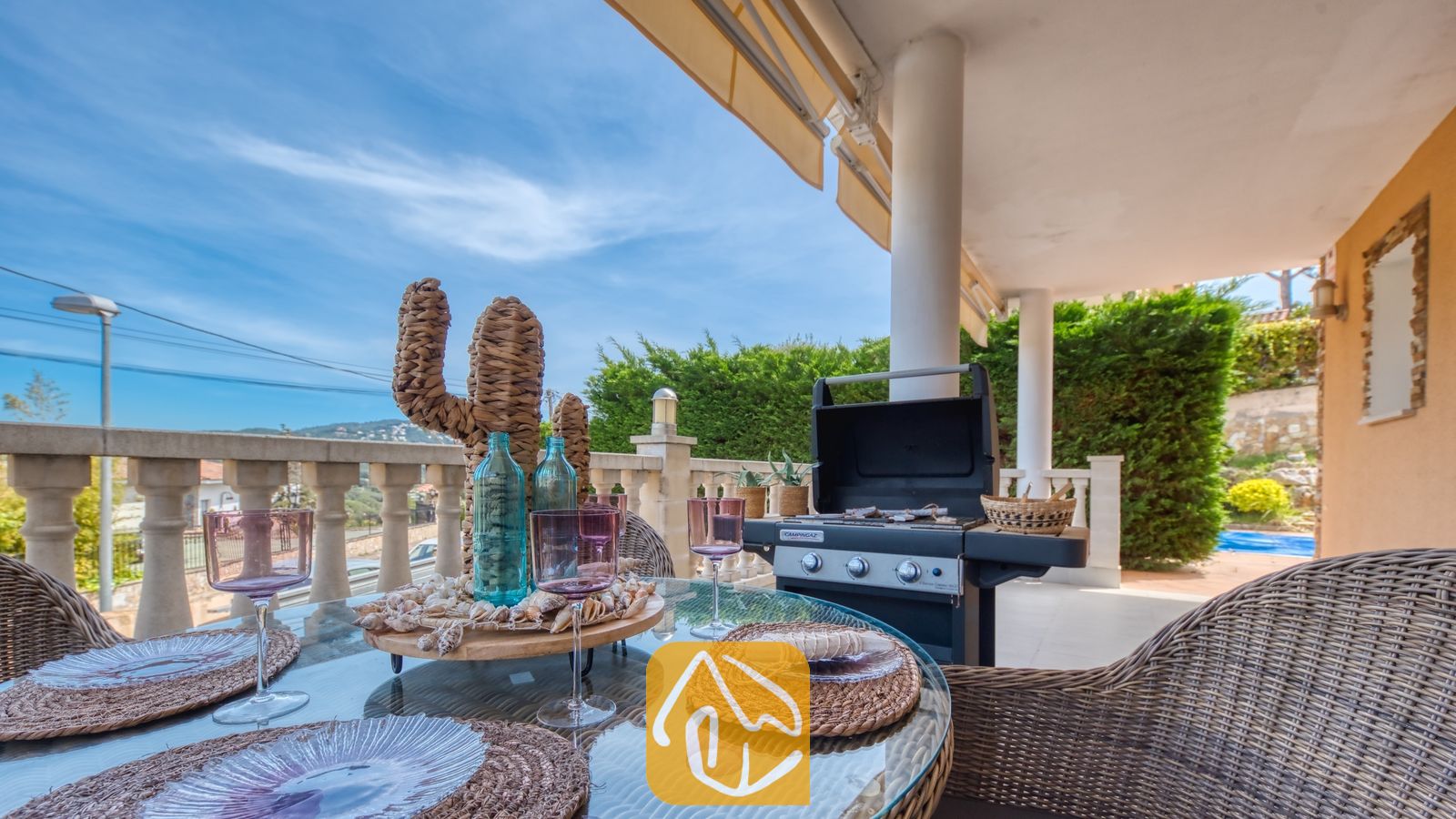 Vakantievilla's Costa Brava Spanje - Villa Primavera - Overdekt terras bij Villa Primavera, voorzien van een glazen eettafel gedekt met geweven placemats, roze wijnglazen en een decoratief rieten cactusmiddelpunt, met een zwarte barbecue in de buurt.