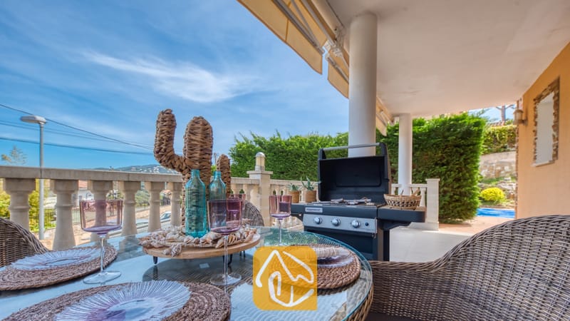 Vakantievilla's Costa Brava Spanje - Villa Primavera - Overdekt terras bij Villa Primavera, voorzien van een glazen eettafel gedekt met geweven placemats, roze wijnglazen en een decoratief rieten cactusmiddelpunt, met een zwarte barbecue in de buurt.