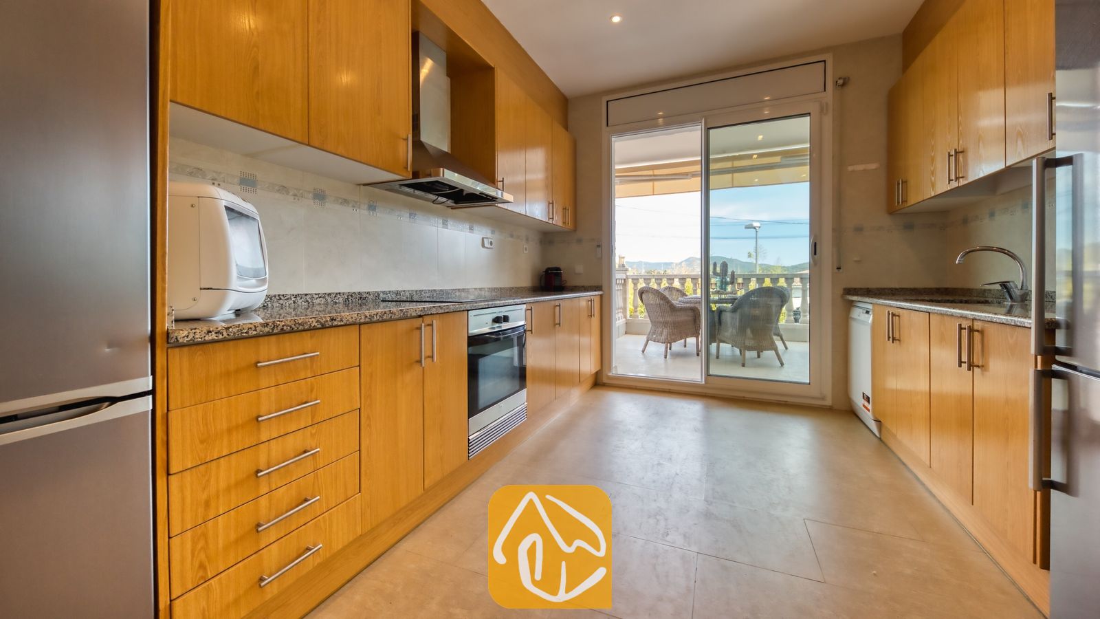 Vakantievilla's Costa Brava Spanje - Villa Primavera - Interieur van de keuken in Villa Primavera met houten kasten, granieten aanrechtbladen en glazen schuifdeuren die uitkomen op een terras.